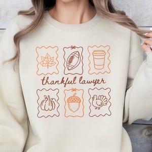 May include: A beige crewneck sweatshirt featuring a graphic print of six rectangular frames containing line art of a maple leaf, football, coffee cup, pumpkin, acorn, and turkey, with the text "thankful lawyer" written in brown script.