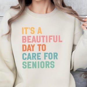 May include: Cream-colored sweatshirt with the text "IT'S A BEAUTIFUL DAY TO CARE FOR SENIORS" in orange, pink, teal, and green. The sweatshirt has long sleeves and a crew neck. The text is centered on the front.