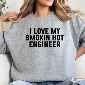 May include: A light grey sweatshirt featuring the text "I LOVE MY SMOKIN HOT ENGINEER" in black. The sweatshirt has a crew neck and long sleeves. The person is wearing blue jeans.