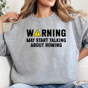 Puede incluir: Sudadera gris claro con el texto "WARNING MAY START TALKING ABOUT ROWING" en negro y amarillo. El símbolo de advertencia es un triángulo amarillo con un signo de exclamación negro. La sudadera es de cuello redondo.