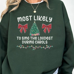 May include: A dark green crewneck sweatshirt featuring the white text "MOST LIKELY TO SING THE LOUDEST DURING CAROLS". The design includes a central green Christmas tree, two red bows on either side, and a string of colourful holiday lights below.
