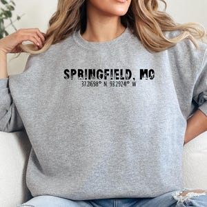 Puede incluir: Sudadera gris claro con el texto negro "SPRINGFIELD, MO" y las coordenadas 37.21698° N, 93.29241° W. La sudadera es de estilo casual, adecuada para el uso diario.