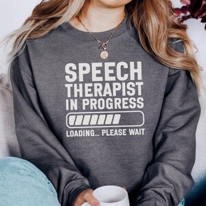 Könnte beinhalten: Dunkelgraues Sweatshirt mit dem weißen Text „SPEECH THERAPIST IN PROGRESS“ und einer Ladebalken-Grafik. Unter der Grafik steht der Satz „LOADING... PLEASE WAIT“. Das Sweatshirt hat einen Rundhalsausschnitt.