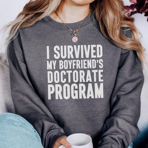 May include: Dark gray sweatshirt with the text "I SURVIVED MY BOYFRIEND'S DOCTORATE PROGRAM" in white block letters. The sweatshirt is worn by a person, and the person is holding a white mug.