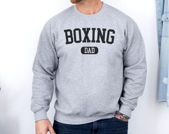 May include: A light gray sweatshirt with the words "BOXING DAD" printed in black block letters. The word "DAD" is in a black rectangle. The sweatshirt is long-sleeved and made of a soft material.