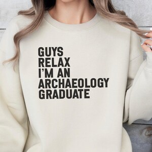 Pode incluir: Uma camisola de gola redonda bege com o texto preto GUYS RELAX I'M AN ARCHAEOLOGY GRADUATE impresso na frente. A peça tem mangas compridas e gola, punhos e bainha canelados, proporcionando um visual simples, limpo e muito casual.