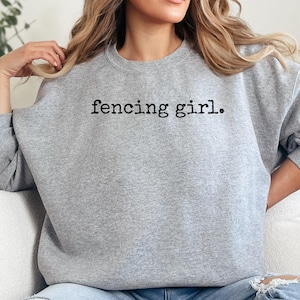 May include: A light gray sweatshirt with the text "fencing girl." in black. The sweatshirt is a crew neck style and has long sleeves. The image shows the sweatshirt on a person.