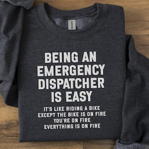 Puede incluir: Sudadera gris oscuro con texto blanco: "SER UN DESPACHADOR DE EMERGENCIAS ES FÁCIL". Debajo, el texto dice: "ES COMO MONTAR EN BICICLETA, EXCEPTO QUE LA BICICLETA ESTÁ EN LLAMAS, TÚ ESTÁS EN LLAMAS, TODO ESTÁ EN LLAMAS".
