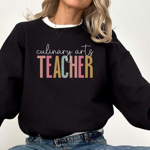 May include: Black sweatshirt with the words "culinary arts TEACHER" in colorful letters. The text is in a script font. The sweatshirt has a white collar and is worn by a person.
