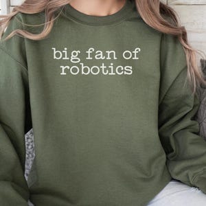 May include: An olive green crewneck sweatshirt featuring the white text "big fan of robotics" printed in a typewriter-style font across the chest. The garment has long sleeves, a ribbed neckline, and a standard fit with a solid color finish.