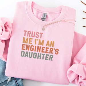Può includere: Una felpa rosa chiaro con la scritta "TRUST ME I'M AN ENGINEER'S DAUGHTER" in un font retrò a blocchi. Una delicata collana d'oro con un piccolo ciondolo è sulla felpa. Le maniche sono piegate.