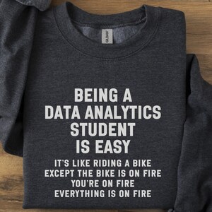 Può includere: Felpa grigio scuro con la scritta bianca: "ESSERE UNO STUDENTE DI DATA ANALYTICS È FACILE". Sotto, il testo continua: "È COME ANDARE IN BICICLETTA TRANNE CHE LA BICI È IN FIAMME, SEI IN FIAMME, TUTTO È IN FIAMME."