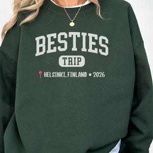 Può includere: Una felpa verde scuro con la scritta "BESTIES TRIP" in lettere bianche. Sotto, "HELSINKI, FINLAND • 2026" con un'icona a forma di puntina rossa. La felpa è a girocollo.