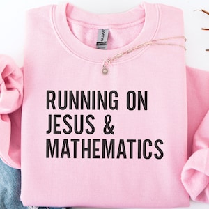 以下が含まれることがあります： 「RUNNING ON JESUS & MATHEMATICS」の文字が黒でプリントされた、明るいピンク色のスウェットシャツ。小さなペンダントが付いた繊細なネックレスがスウェットシャツの上に置かれています。袖は折りたたまれ、青いデニムの一部が見えています。