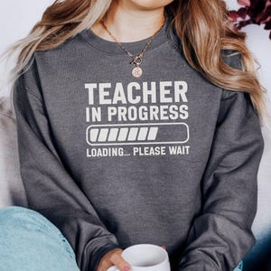 Może przedstawiać: Ciemnoszara bluza z białym napisem „TEACHER IN PROGRESS”, graficznym paskiem ładowania i słowami „LOADING... PLEASE WAIT”. Bluza ma swobodny styl.