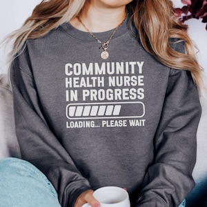 Peut inclure: Sweat-shirt gris foncé avec l'inscription blanche « COMMUNITY HEALTH NURSE IN PROGRESS ». En dessous, un graphique de barre de chargement et les mots « LOADING... PLEASE WAIT ». Le sweat-shirt est de style col rond.