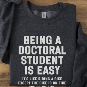 May include: A white ceramic mug with a black handle and the text "But First, Coffee" in a black, bold font. The mug is on a white surface. The mug is approximately 10 cm tall.