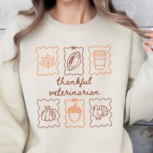 Puede incluir: Una sudadera de cuello redondo color beige con un estampado gráfico de seis marcos rectangulares estilo sello postal que contienen dibujos lineales de una hoja de arce, un balón de fútbol americano, un vaso de café, una calabaza, una bellota y un pavo.