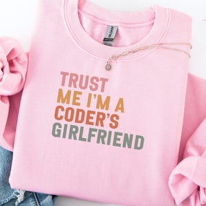 Könnte beinhalten: Hellrosa Sweatshirt mit dem Text "TRUST ME I'M A CODER'S GIRLFRIEND" in Orange, Braun und Grün. Eine Halskette mit einem Sonnenanhänger liegt auf dem Sweatshirt.