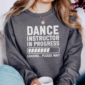 Może przedstawiać: Ciemnoszara bluza z białym napisem "DANCE INSTRUCTOR IN PROGRESS" i grafiką paska ładowania. Poniżej grafiki znajduje się fraza "LOADING... PLEASE WAIT". Bluza ma swobodny styl.