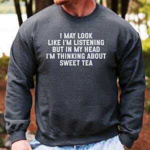 May include: A dark grey crew neck sweatshirt with the white text: "I MAY LOOK LIKE I'M LISTENING BUT IN MY HEAD I'M THINKING ABOUT SWEET TEA". The sweatshirt is made of a soft material. The background is a blurred outdoor scene.