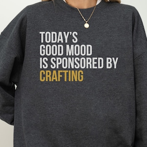 以下が含まれることがあります： ダークチャコールグレーのクルーネックトレーナー。白い文字で「TODAY'S GOOD MOOD IS SPONSORED BY」、マスタードイエローの文字で「CRAFTING」とプリントされています。長袖でリブ編みの襟元が特徴の、シンプルでカジュアルなデザインの衣類です。