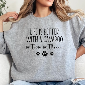 May include: A light gray sweatshirt with the text "LIFE IS BETTER WITH A CAVAPOO or two, or three..." in black. Below the text are three black paw prints. The sweatshirt is a crew neck style.