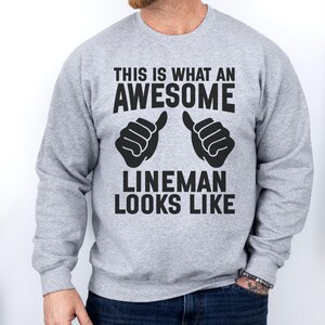 Op de afbeelding: Een lichtgrijs sweatshirt met de tekst "THIS IS WHAT AN AWESOME LINEMAN LOOKS LIKE" in het zwart. Het ontwerp bevat twee duim omhoog gebaren. De sweatshirt is een crew neck stijl.