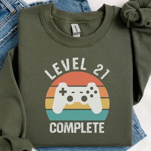 May include: Olive green sweatshirt with a retro-style graphic. The graphic features a white game controller inside a colorful sunset design, with the text "LEVEL 21 COMPLETE" in white. The sweatshirt is folded on top of a pair of blue jeans.
