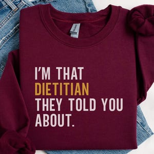 May include: Burgundy sweatshirt with the text "I'M THAT DIETITIAN THEY TOLD YOU ABOUT." in white and gold lettering. The sweatshirt is folded on top of a pair of blue jeans.