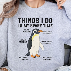 May include: A light gray crewneck sweatshirt featuring a central graphic of a penguin. The text reads: THINGS I DO IN MY SPARE TIME, with labels surrounding the penguin: WATCH PENGUINS, RESEARCH PENGUINS, LOOK AT PENGUINS, DREAM ABOUT PENGUINS, TALK ABOUT PENGUINS, THINK ABOUT PENGUINS.