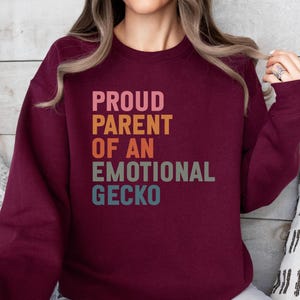 Może przedstawiać: Bordowa bluza z napisem "PROUD PARENT OF AN EMOTIONAL GECKO" w kolorowej, ułożonej czcionce. Tekst jest w odcieniach różu, pomarańczy, turkusu i niebieskiego. Bluza jest w jednolitym kolorze i ma okrągły dekolt.