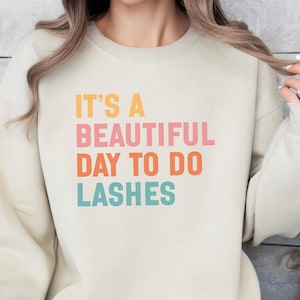 May include: A cream-colored sweatshirt with the text "IT'S A BEAUTIFUL DAY TO DO LASHES" in orange, pink, orange, and teal lettering. The sweatshirt has long sleeves and a crew neck.