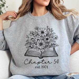 May include: A gray sweatshirt featuring a floral bouquet design atop an open book. The text "Chapter 54 est. 1971" is printed below the book. The sweatshirt is a crew neck style.
