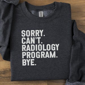 May include: Dark gray sweatshirt with the text "SORRY. CAN'T. RADIOLOGY PROGRAM. BYE." in white block letters. The sweatshirt is folded on a wooden surface. The Gildan brand tag is visible.