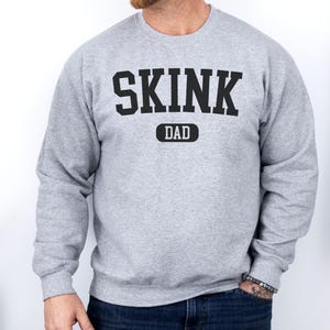 May include: A grey sweatshirt featuring the text "SKINK DAD" in large, black, block letters. The word "DAD" is in a smaller, black, rectangular box below "SKINK". The sweatshirt has long sleeves.