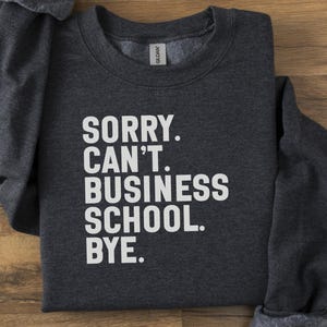 Puede incluir: Sudadera gris oscuro con el texto blanco "SORRY. CAN'T. BUSINESS SCHOOL. BYE.". La prenda está doblada sobre una superficie de madera.