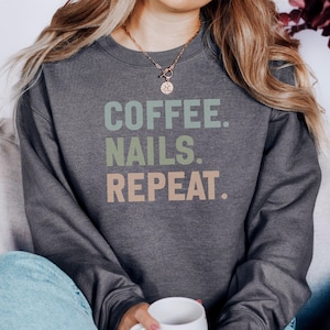 May include: Dark grey crew neck sweatshirt with the text "COFFEE. NAILS. REPEAT." in green and peach. The sweatshirt is worn by a person. A gold necklace is visible. The person is holding a white mug.