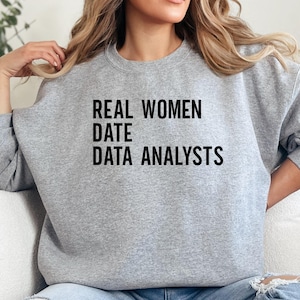 May include: A light gray sweatshirt with the text "REAL WOMEN DATE DATA ANALYSTS" in black block letters. The sweatshirt is a crew neck style and appears to be made of a soft material. The overall theme is related to data science and women.