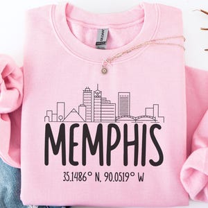 May include: A light pink sweatshirt with the word "MEMPHIS" in large black letters. Above the word is a black line drawing of the Memphis skyline. The coordinates 35.1486° N, 90.0519° W are printed below the word. A necklace with a small pendant rests on the sweatshirt.