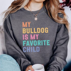 Puede incluir: Sudadera gris oscuro con el texto "MY BULLDOG IS MY FAVORITE CHILD" en naranja, amarillo, rosa, turquesa y morado. La sudadera es de cuello redondo.