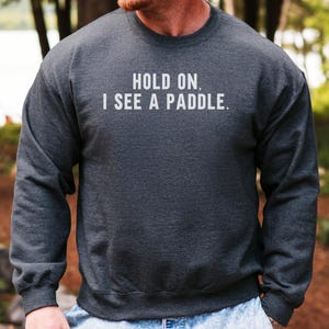 May include: Dark grey sweatshirt with the text "HOLD ON, I SEE A PADDLE." The long-sleeved crew neck shirt is made of a soft material. The background shows a blurred outdoor scene with trees and water.