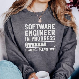 Könnte beinhalten: Dunkelgraues Sweatshirt mit dem weißen Text "SOFTWARE ENGINEER IN PROGRESS", einer Ladebalken-Grafik und den Worten "LOADING... PLEASE WAIT". Das Sweatshirt hat einen Rundhalsausschnitt.
