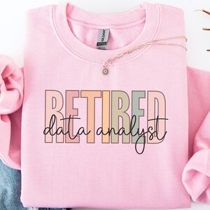 May include: A light pink crewneck sweatshirt featuring the word RETIRED in large, blocky, multicolored pastel letters, with the words data analyst written in a black cursive script font layered over the center of the text.
