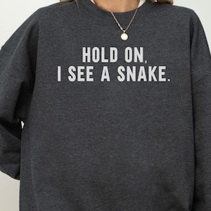 May include: A dark charcoal grey crewneck sweatshirt featuring the white text "HOLD ON, I SEE A SNAKE." printed across the chest. The garment has long sleeves and a ribbed neckline, providing a simple and straightforward design for casual wear.