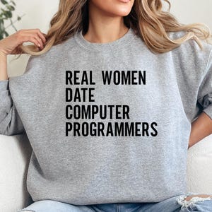 以下が含まれることがあります： 「REAL WOMEN DATE COMPUTER PROGRAMMERS」の黒い文字がプリントされた、ヘザーグレーのスウェットシャツ。カジュアルなスタイルで、普段使いに適しています。