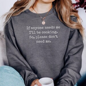 Könnte beinhalten: Dunkelgraues Sweatshirt mit dem Text: „Wenn mich jemand braucht, koche ich. Also, bitte, braucht mich nicht.“ Das Sweatshirt hat einen Rundhalsausschnitt. Die Person trägt eine blaue Jeans und hält eine weiße Tasse.