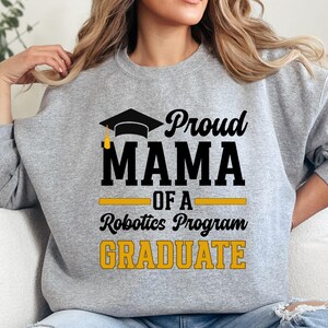 May include: A gray sweatshirt with the text "Proud MAMA of a Robotics Program GRADUATE" in black and yellow. The design includes a graduation cap. The sweatshirt is a comfortable, casual style.