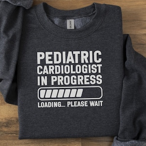 Könnte beinhalten: Dunkelgraues Sweatshirt mit weißem Text, der "Pediatric Cardiologist In Progress" über einer Ladebalken-Grafik und den Worten "Loading... Please Wait" zeigt. Das Sweatshirt liegt gefaltet auf einer Holzoberfläche.