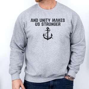 Puede incluir: Una sudadera gris claro con el texto "AND UNITY MAKES US STRONGER" sobre un gráfico de ancla negra. La sudadera es de manga larga y tiene cuello redondo.